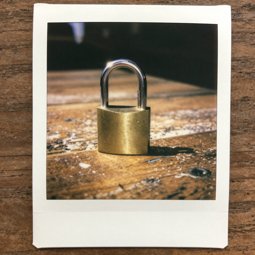 instant Polaroid photograph, vintage 1970s aesthetic, faded colors, white border frame, slightly overexposed, nostalgic lo-fi quality, amateur snapshot, a small steel padlock, half-open mid-snap with spring tension visible, resting on a weathered wooden table, sunlight from the side catching the curved shackle as it closes, atmosphere of quiet finality [Z-Image Turbo]
