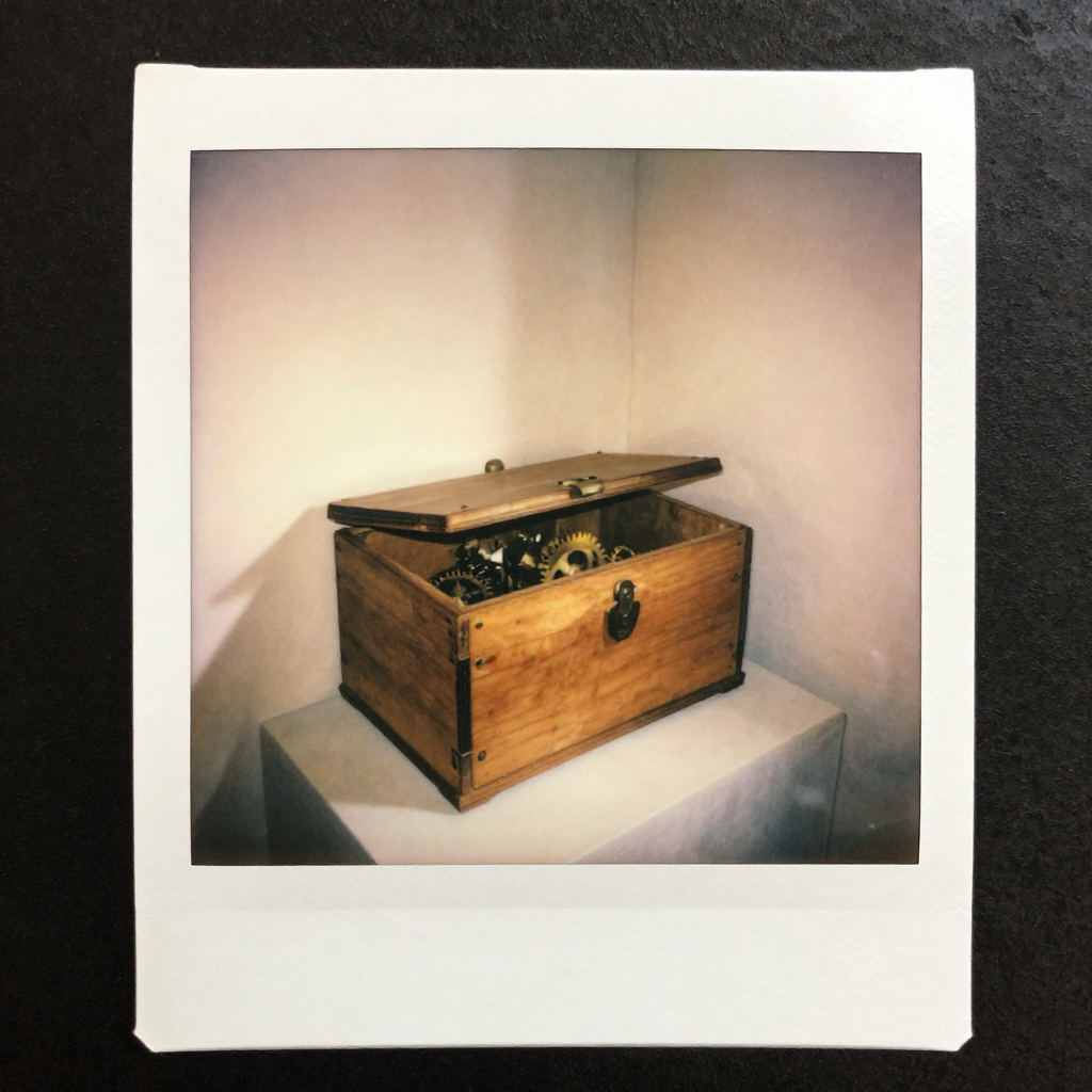instant Polaroid photograph, vintage 1970s aesthetic, faded colors, white border frame, slightly overexposed, nostalgic lo-fi quality, amateur snapshot, a small wooden lockbox sitting slightly off-center on a sunlit wall corner, its surface worn but warm, a faint golden light pulsing from within as intricate gears silently shift and reconfigure behind the locked lid, morning light from the left casting a soft shadow, stillness and quiet transformation in the air [Z-Image Turbo]