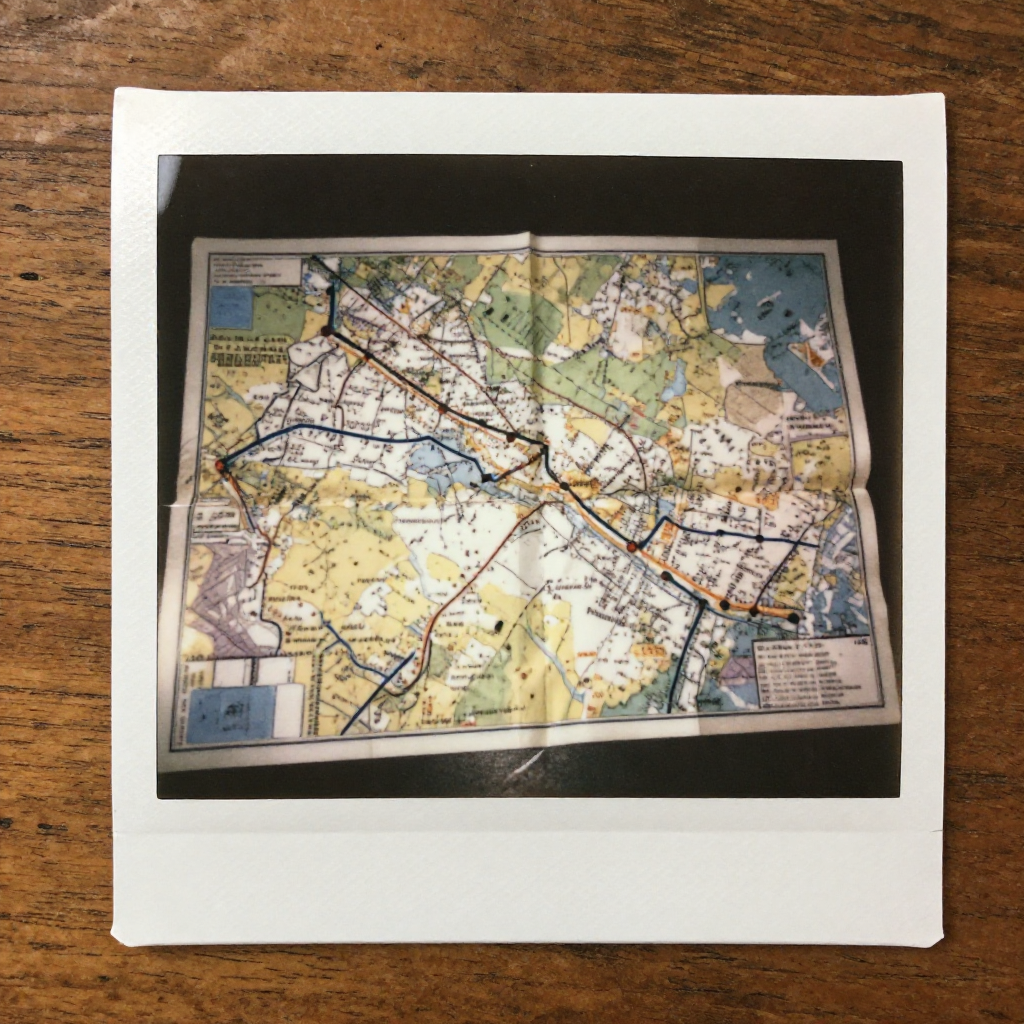 instant Polaroid photograph, vintage 1970s aesthetic, faded colors, white border frame, slightly overexposed, nostalgic lo-fi quality, amateur snapshot, A crumpled paper map lying on a worn wooden table, its surface dusted with old coffee rings and faint pencil smudges, sunlight from a window to the left casting soft shadows across hand-drawn route corrections that don’t align with printed roads, one path subtly rewritten in different ink, barely noticeable, the paper torn along a hidden fold where the real destination used to be [Z-Image Turbo]