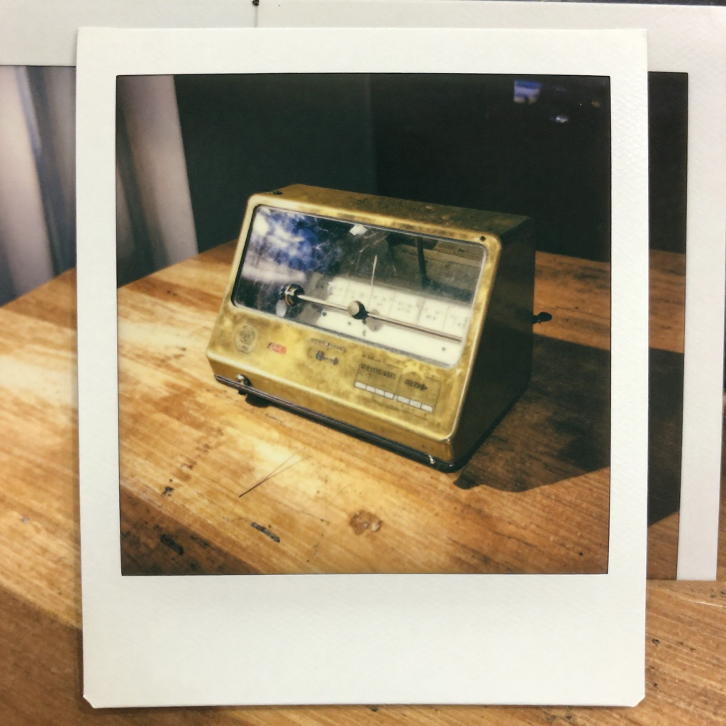 instant Polaroid photograph, vintage 1970s aesthetic, faded colors, white border frame, slightly overexposed, nostalgic lo-fi quality, amateur snapshot, a salvaged mirror galvanometer, brass casing with faint tarnish and glass enclosure cracked at one edge, sunlight from the left casting a delicate needle shadow across a worn pine table, stillness heavy with forgotten urgency [Z-Image Turbo]